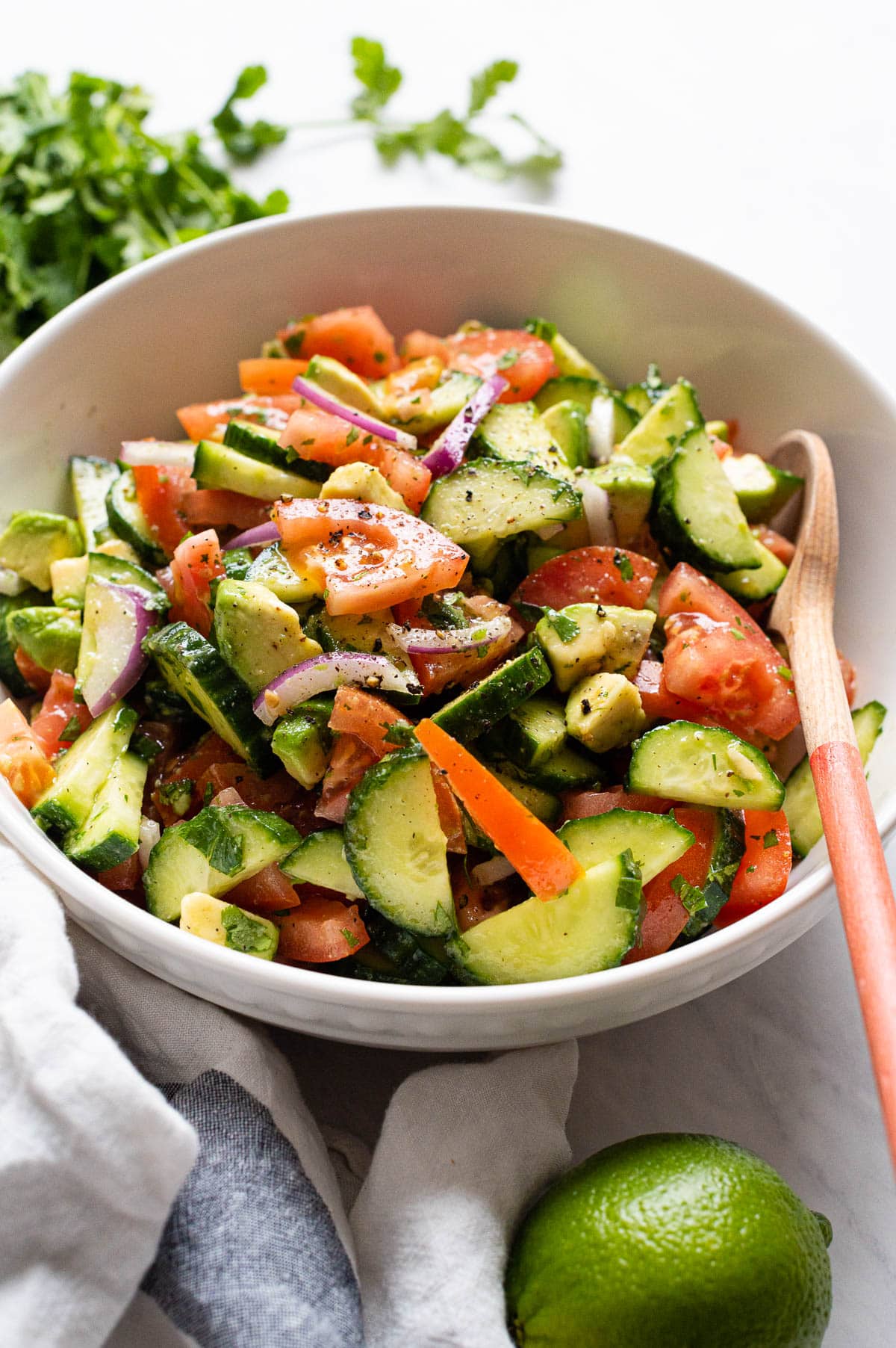 Cucumber tomato avocado salad served in a bowl with a wooden spoon and a bunch of cilantro, lime and linen towel on a counter.