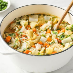 White pot of chicken noodle vegetable soup and wooden spoon in it.