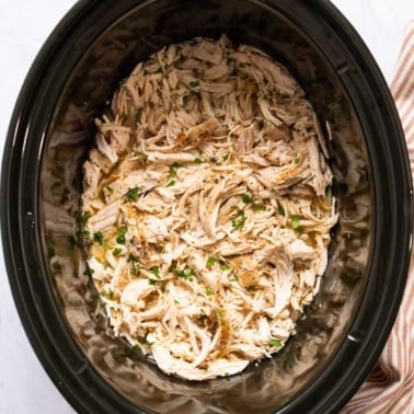 Shredded chicken in broth garnished with herbs in black slow cooker.