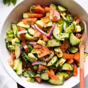 Cucumber tomato avocado salad in white bowl with wooden spoon.