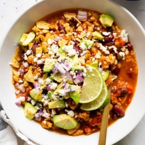 Ground chicken chili served with avocado, red onion, cheese and lime in white bowl with a spoon.