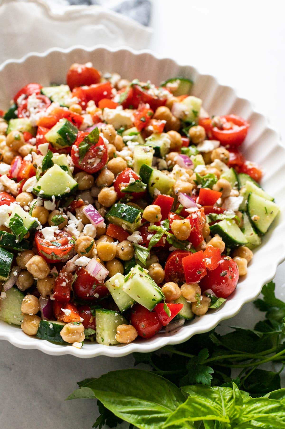 Side view of Mediterranean salad with chickpeas served in white bowl and fresh herbs nearby.