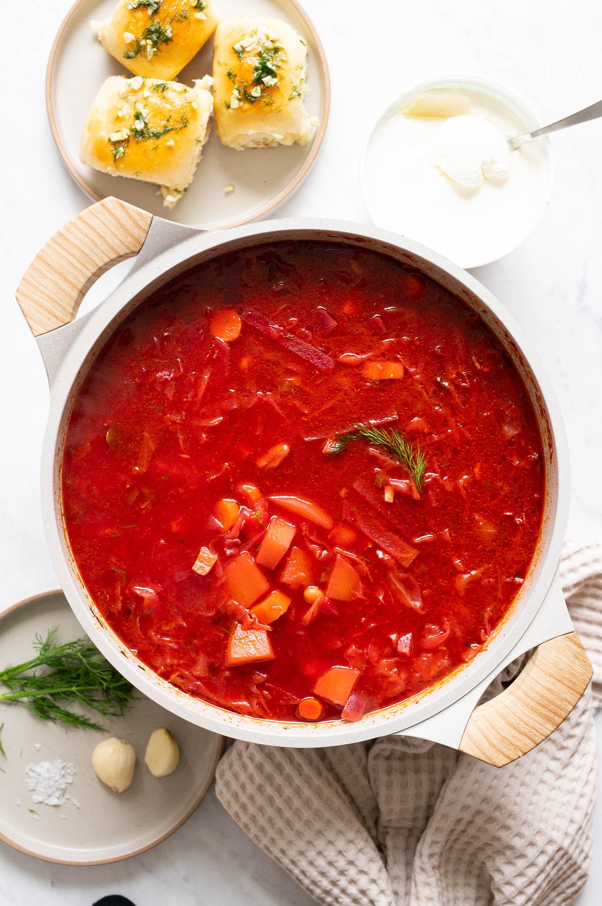 A pot with borsch and pampushky, sour cream, dill and garlic served on a side.