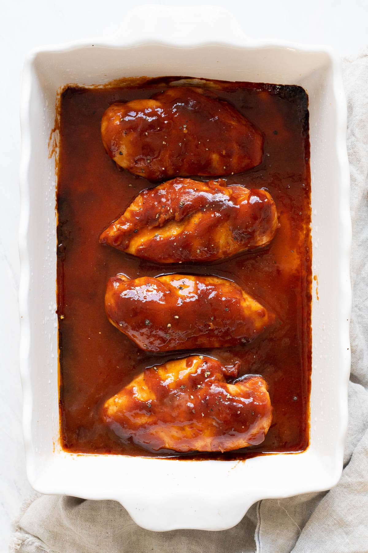 Four BBQ chicken breasts in white baking dish and linen on a counter.