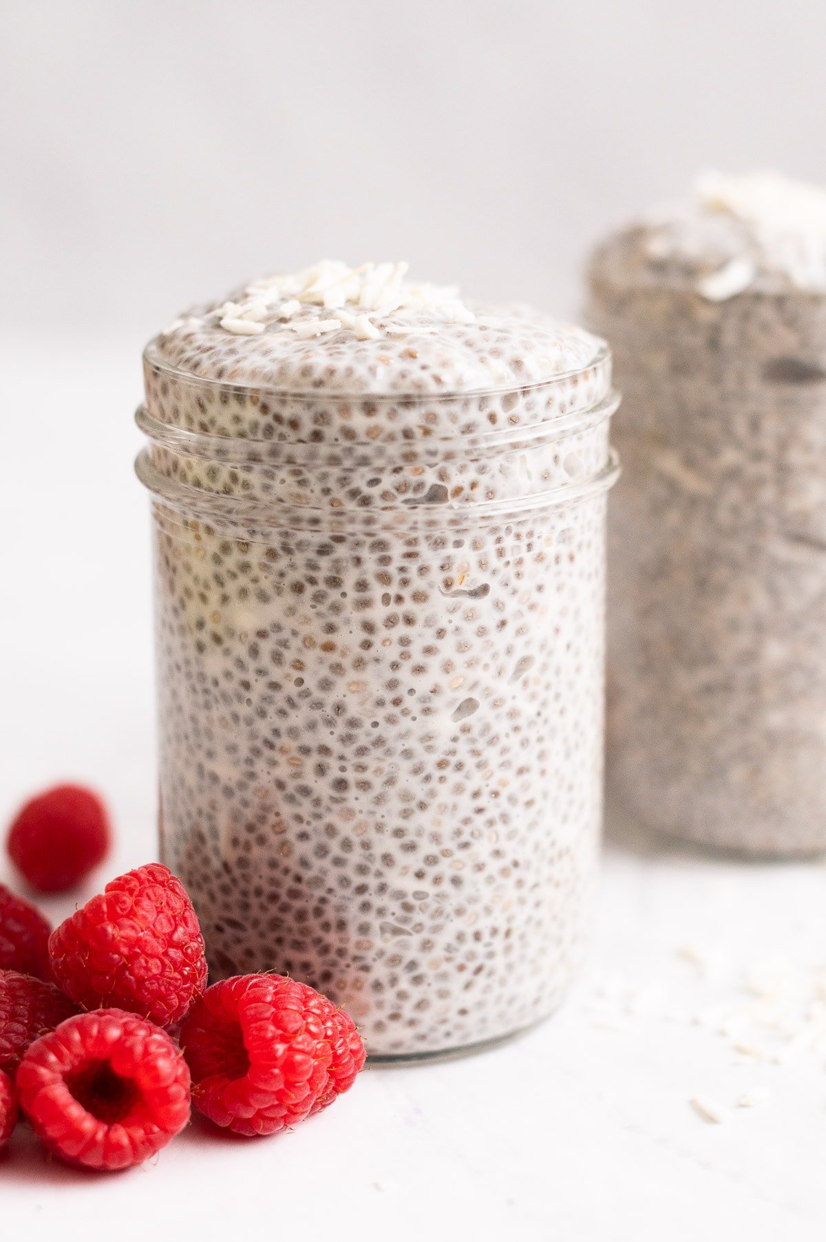 Coconut milk chia pudding topped with coconut flakes in mason jars and fresh raspberries on a counter.
