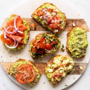 Six avocado toasts with various toppings on a serving board.