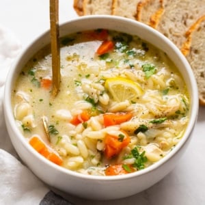 A bowl of lemon chicken orzo soup with a spoon and bread next to it.