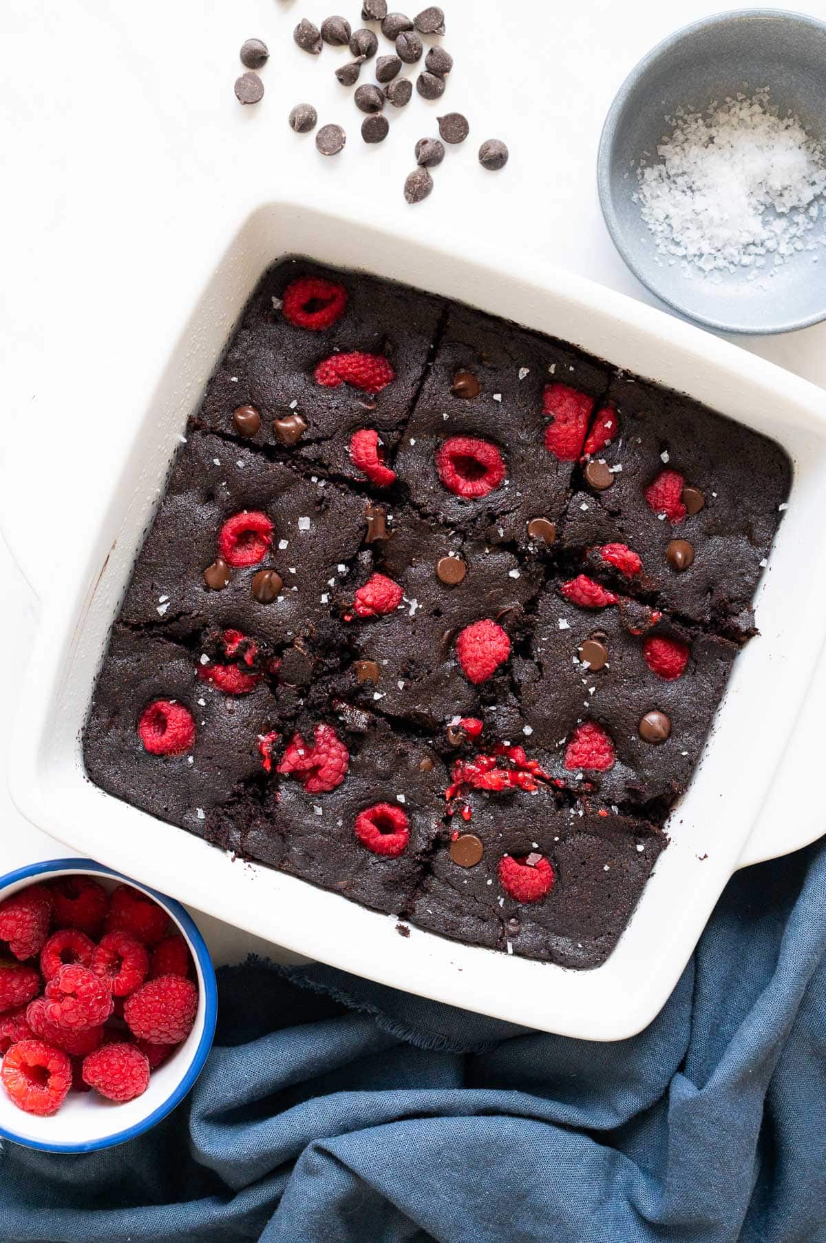 Sliced oat flour brownies with chocolate chips and raspberries in white baking dish.