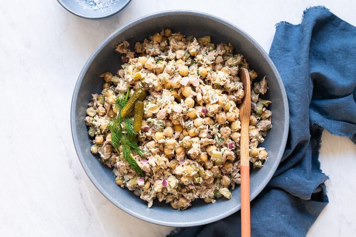 Chickpea salad with tuna garnished with pickles and dill served in a bowl with linen towel near on a countertop.