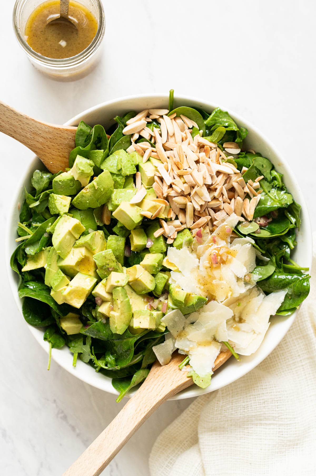 Spinach avocados salad drizzled with dressing in white bowl with tongs.
