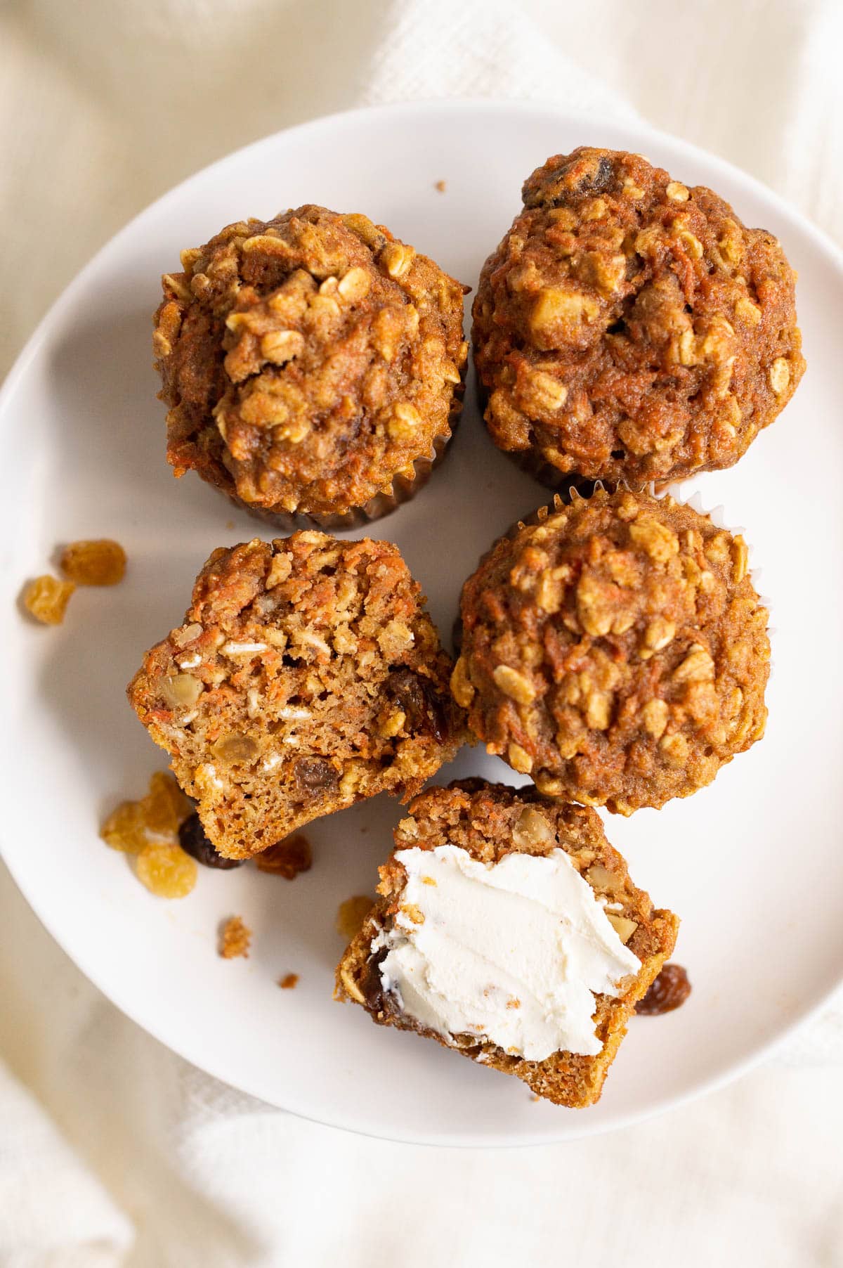 Healthy carrot muffin cut in half and spread with cream cheese and more muffins and raisins on a plate.