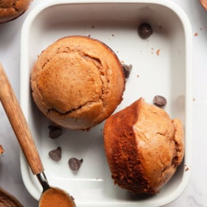 Two protein muffins in a baking dish with chocolate chips and spoonful of nut butter.