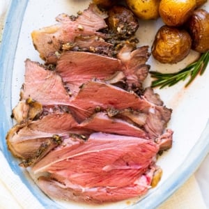 Sliced roast leg of lamb served on a platter with baby potatoes and rosemary.