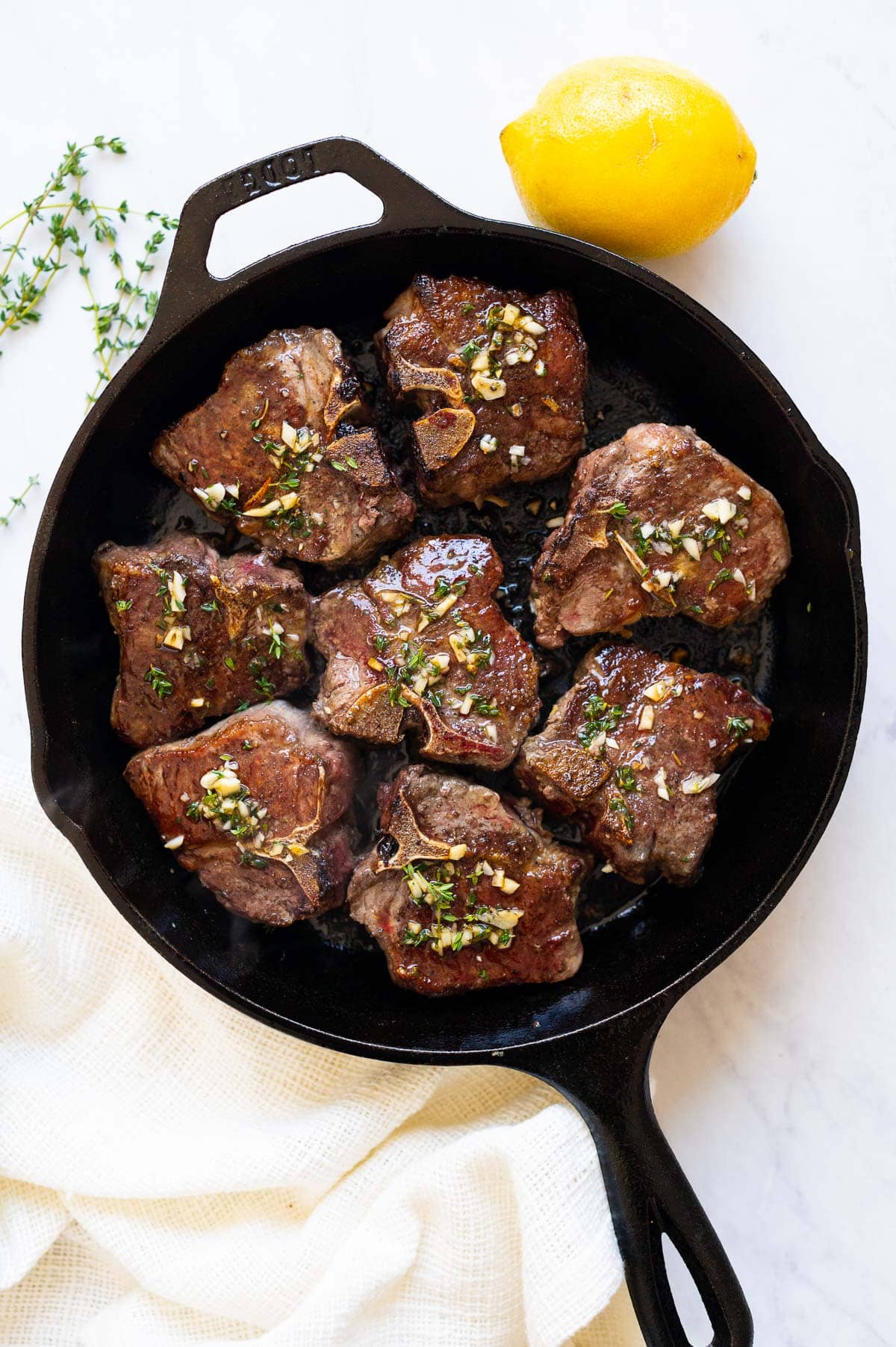 Eight pan seared lamb loin chops in cast iron skillet and napkin, lemon and fresh thyme on a counter.