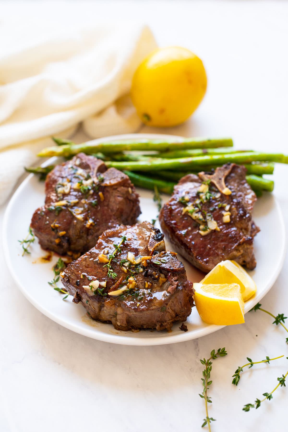 Three lamb loin chops garnished with garlic and fresh thyme served on a plate with asparagus and lemon slices.