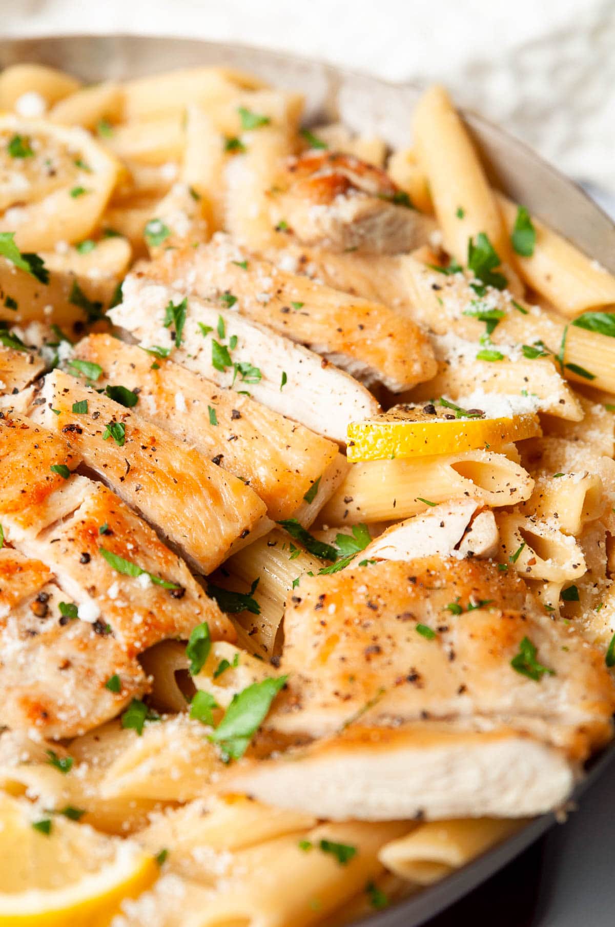 Sliced lemon chicken breast with pasta and lemons garnished with Parmesan and parsley.