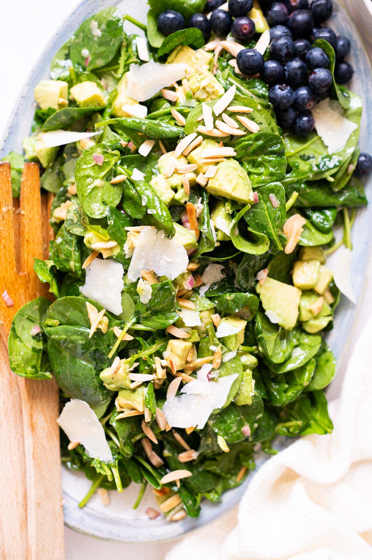 Spinach avocado salad with Parmesan shavings and toasted almonds served on a platter with wooden spoons.