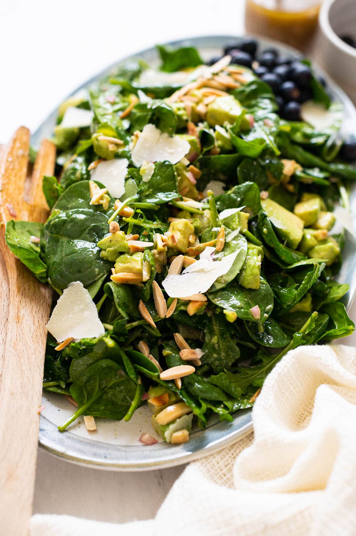Spinach salad with avocado, almonds, parmesan cheese on a serving platter with wooden tongs.