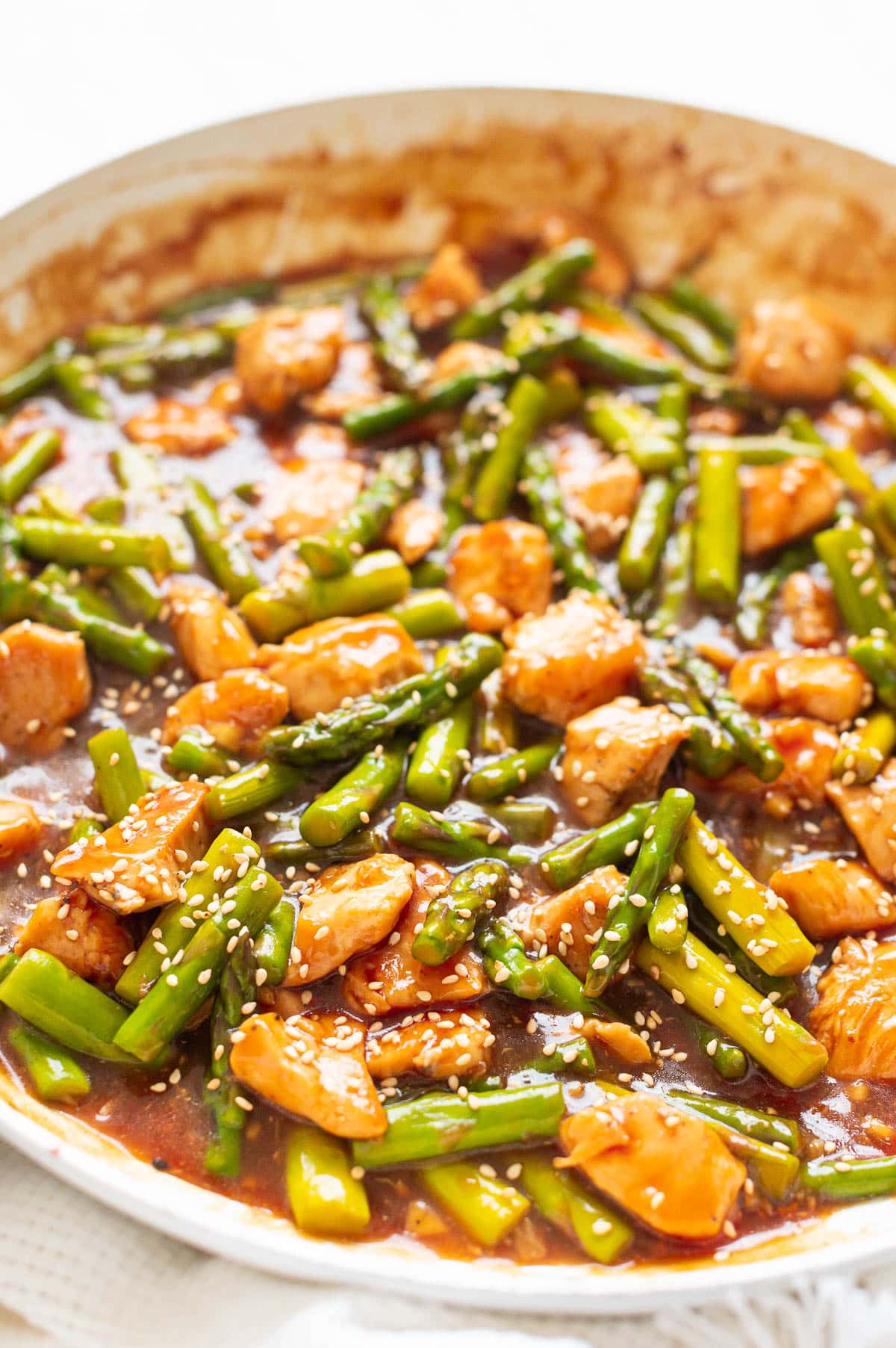 Close up of chicken and asparagus stir fry covered in sauce and sesame seeds in white skillet.