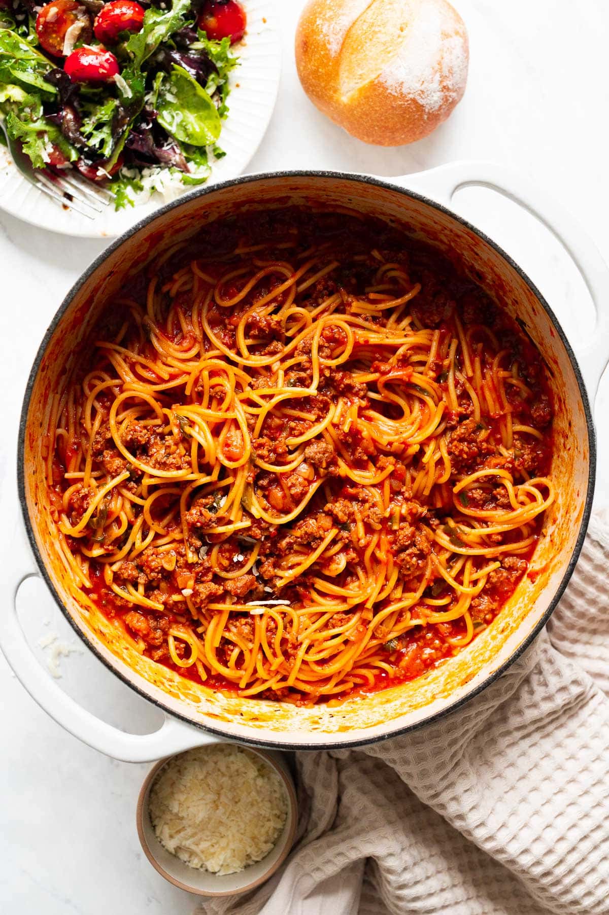 One pot spaghetti recipe served in white pot and salad, bun and parmesan cheese on a counter.