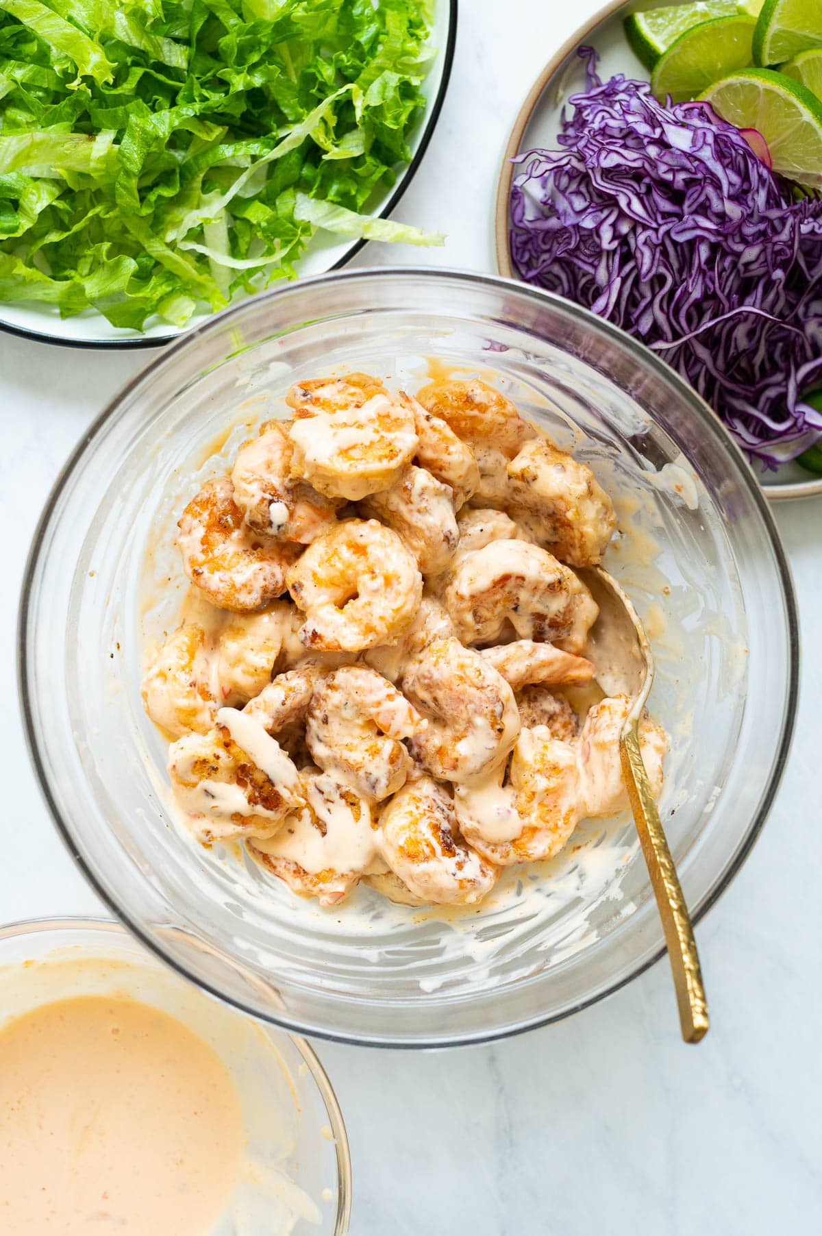 Bang bang shrimp in a bowl with a spoon and lettuce and other toppings around it.