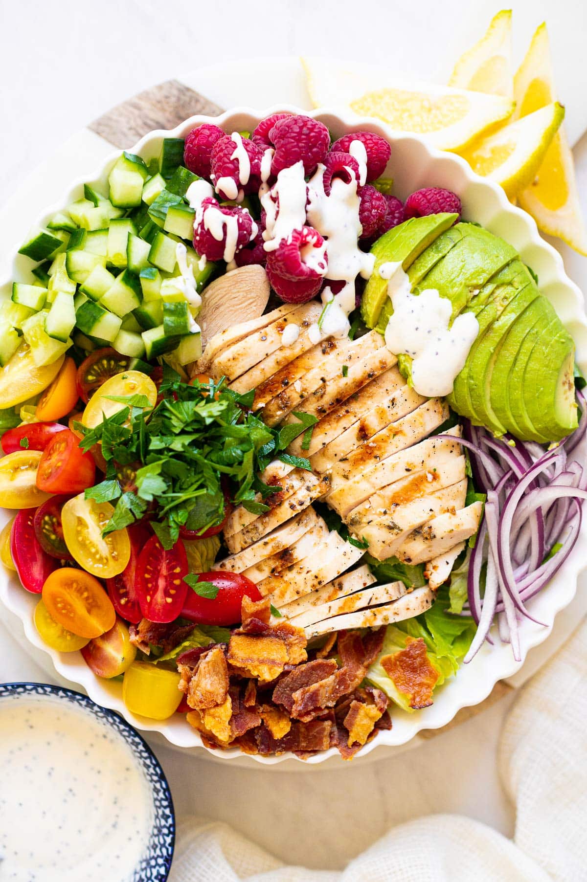 A bowl with salad with grilled chicken, avocado, red onion, parsley, lettuce and bacon. Lemon slices and salad dressing on the side.