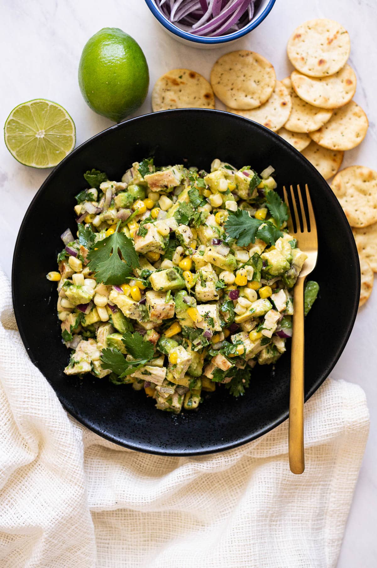 Chicken salad with avocado, corn, cilantro and red onion in black bowl and crackers and lime on a counter.