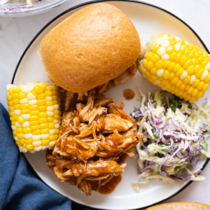Crockpot BBQ pulled chicken served on a bun with coleslaw and corn on a plate.