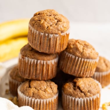Five vegan banana muffins stacked on a plate and bananas behind.