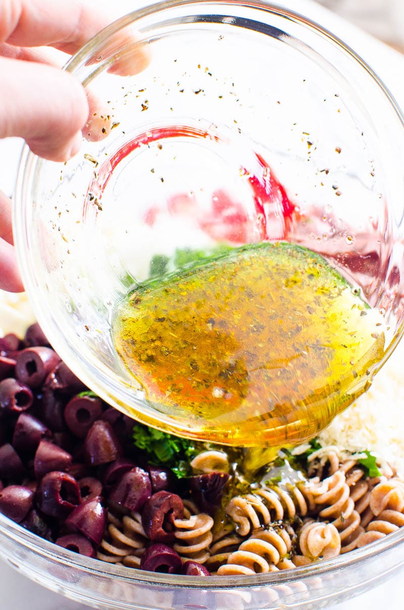 Person pouring Italian salad dressing over pasta salad ingredients in a bowl.