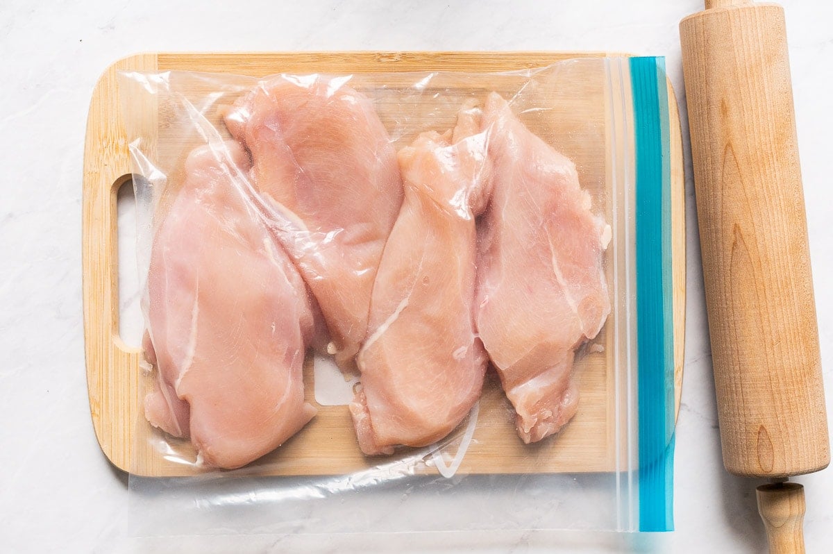 Fresh chicken breasts inside a plastic bag on a cutting board and rolling pin nearby.