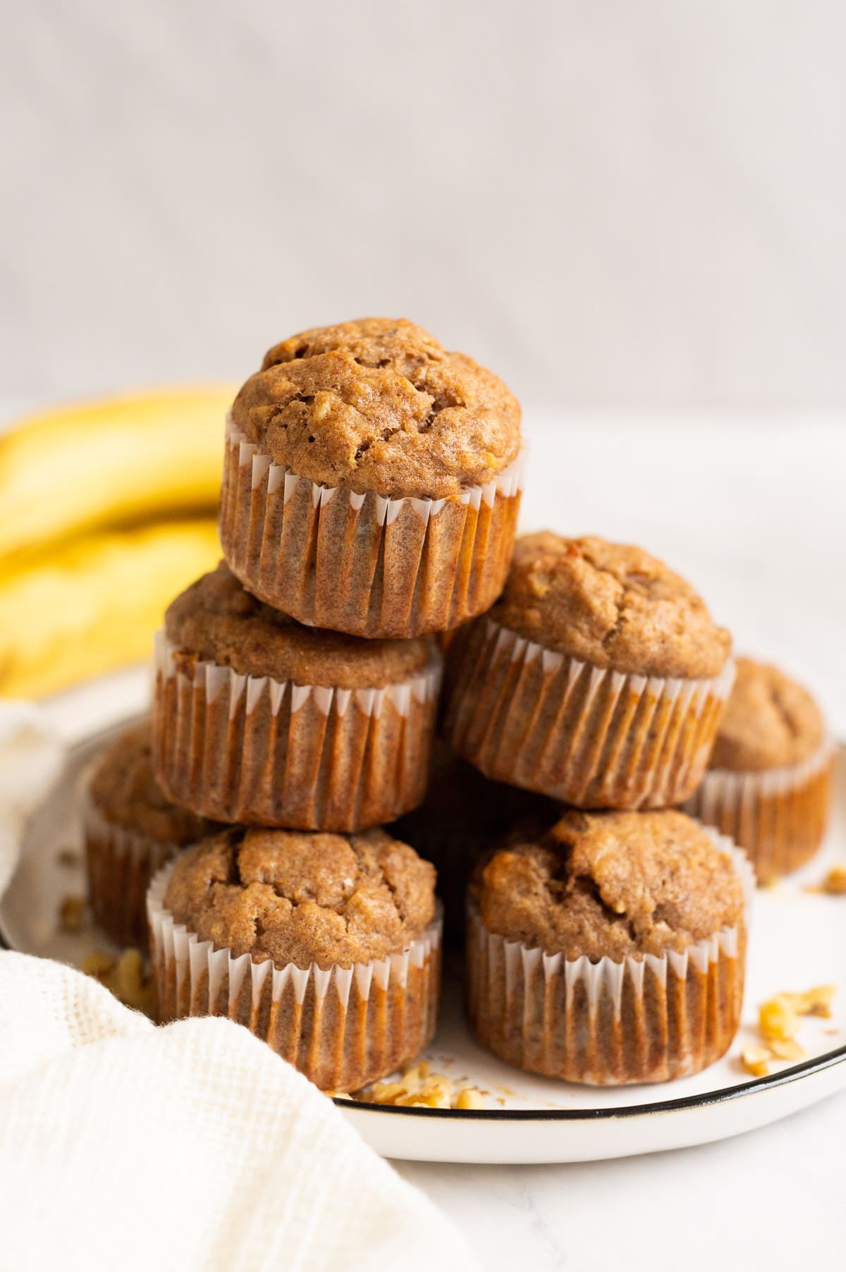 Five vegan banana muffins stacked on a plate and bananas behind.