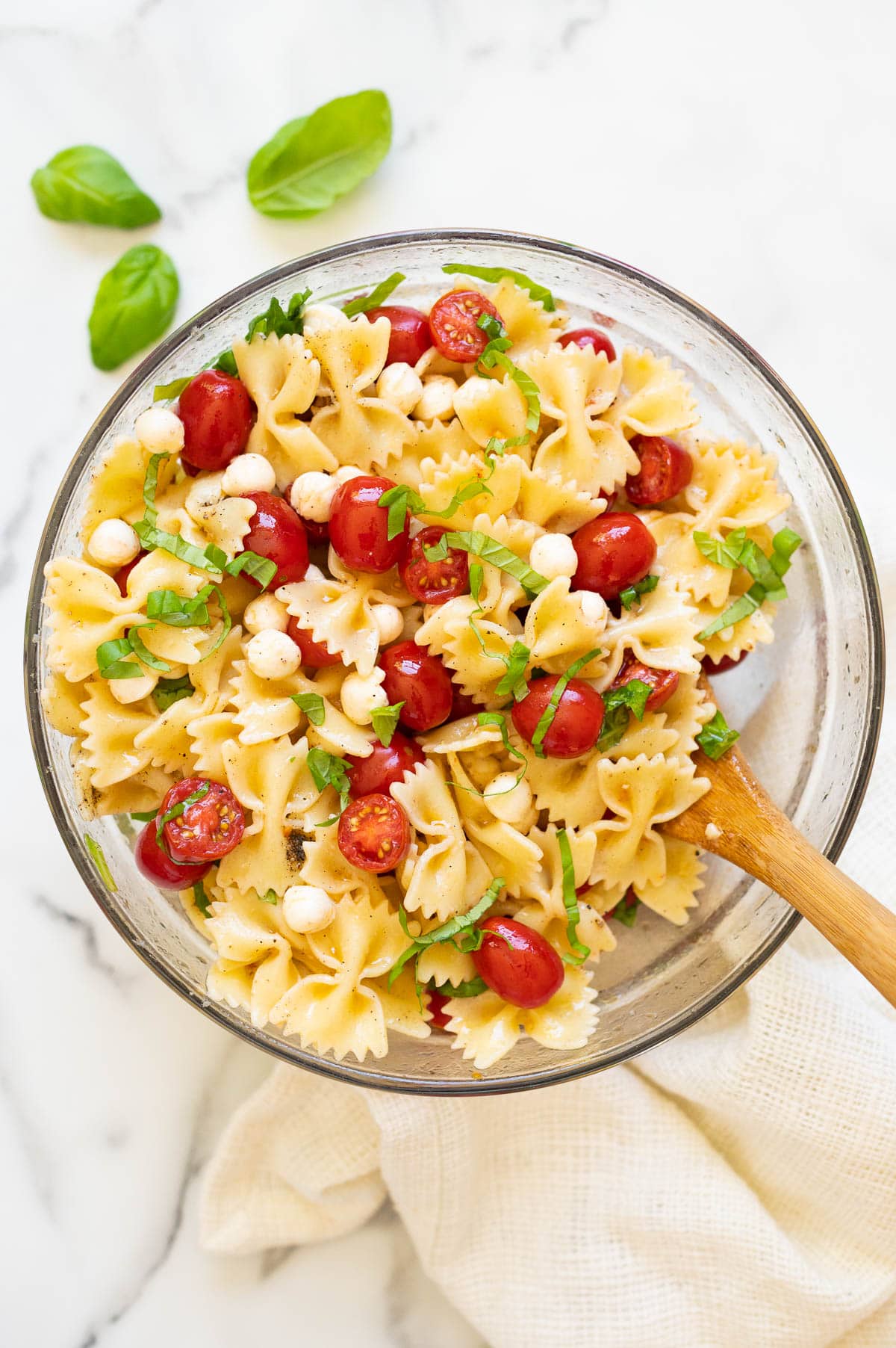 Caprese pasta salad garnished with fresh basil in a glass bowl with wooden spoon.