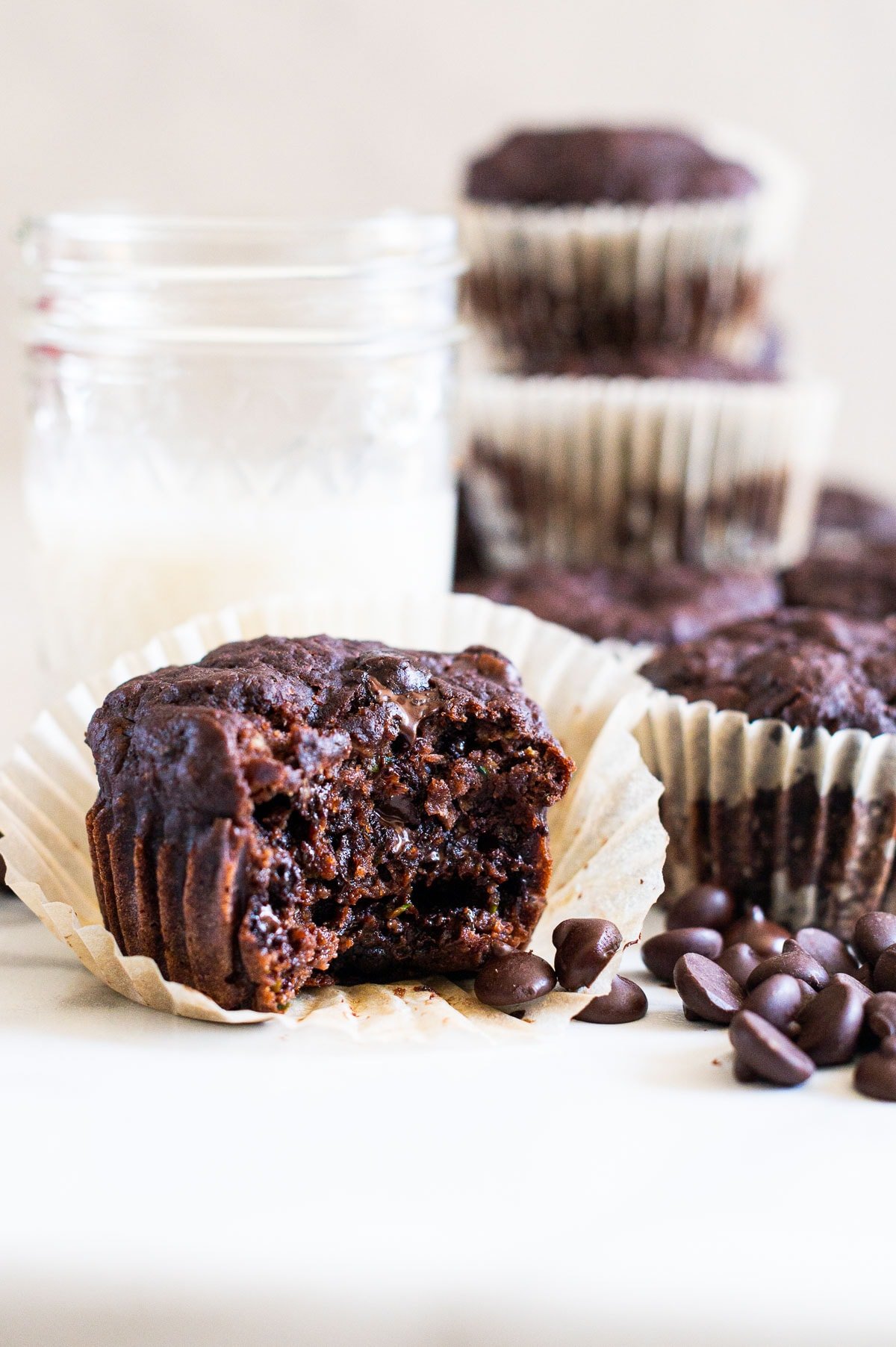 Chocolate zucchini muffins on a countertop with more chocolate chips and a glass of milk.