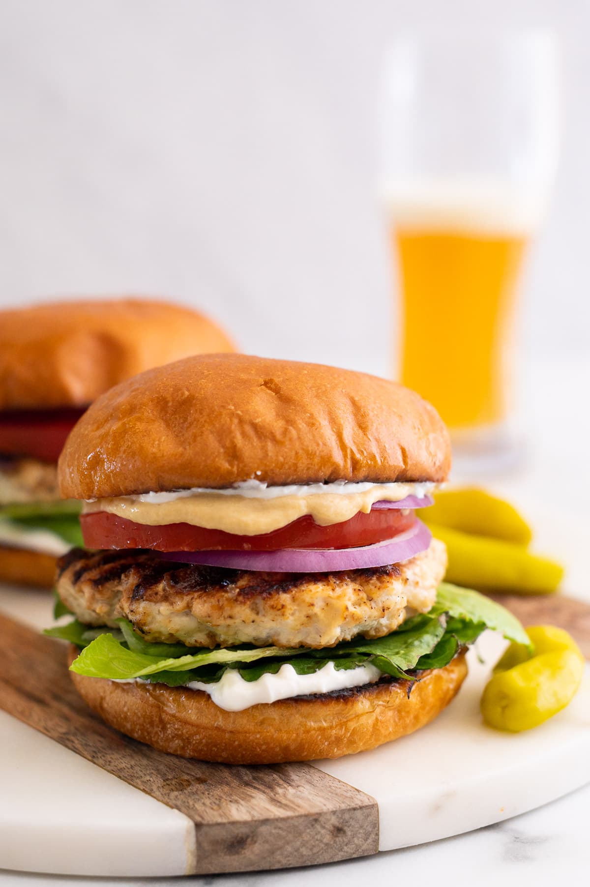 Grilled chicken burgers on a platter with beer in a glass and peperoncini on a side.