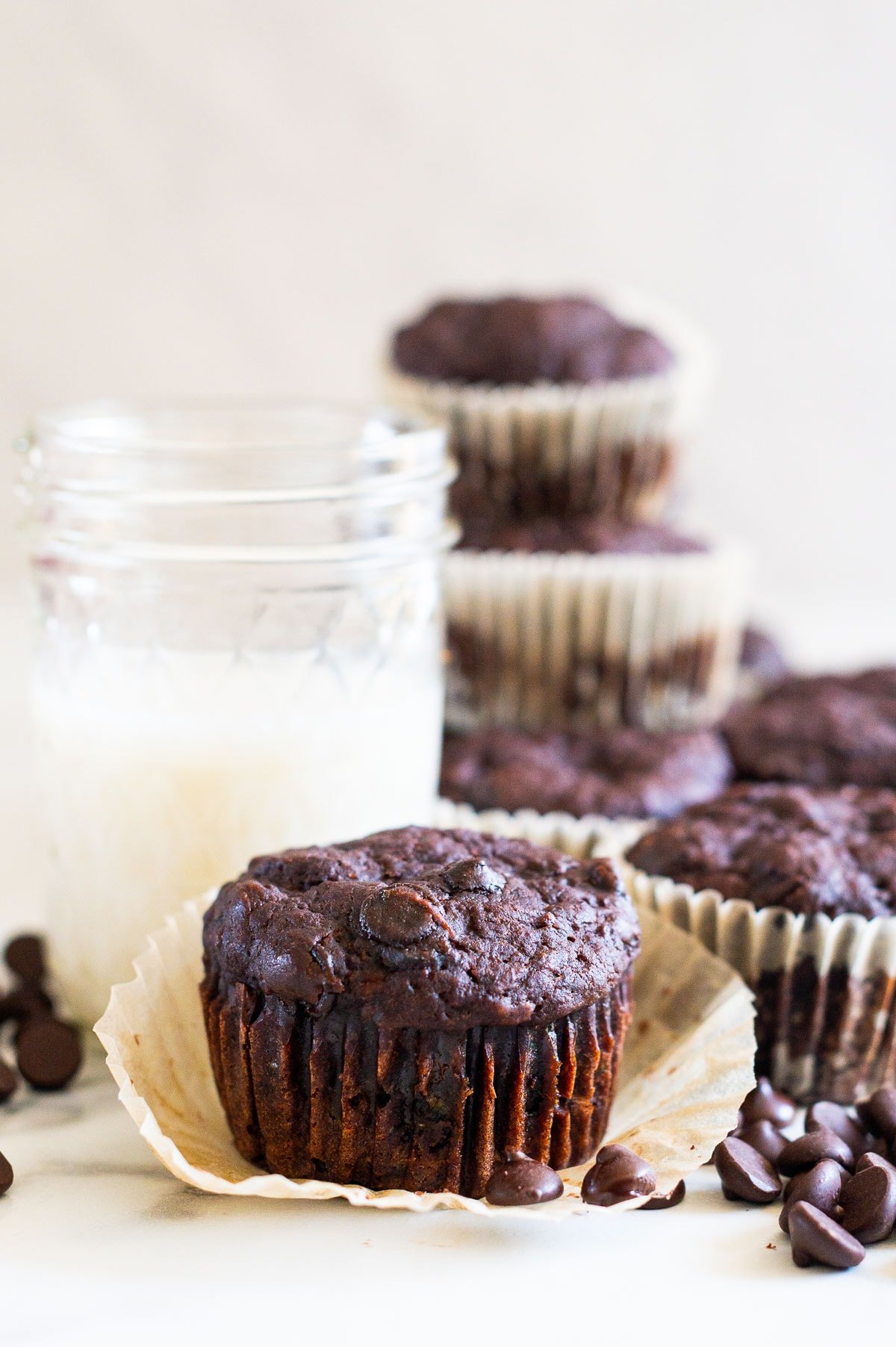 Zucchini chocolate muffin in a wrapper on a countertop surrounded by chocolate chips, muffins and a glass of milk.