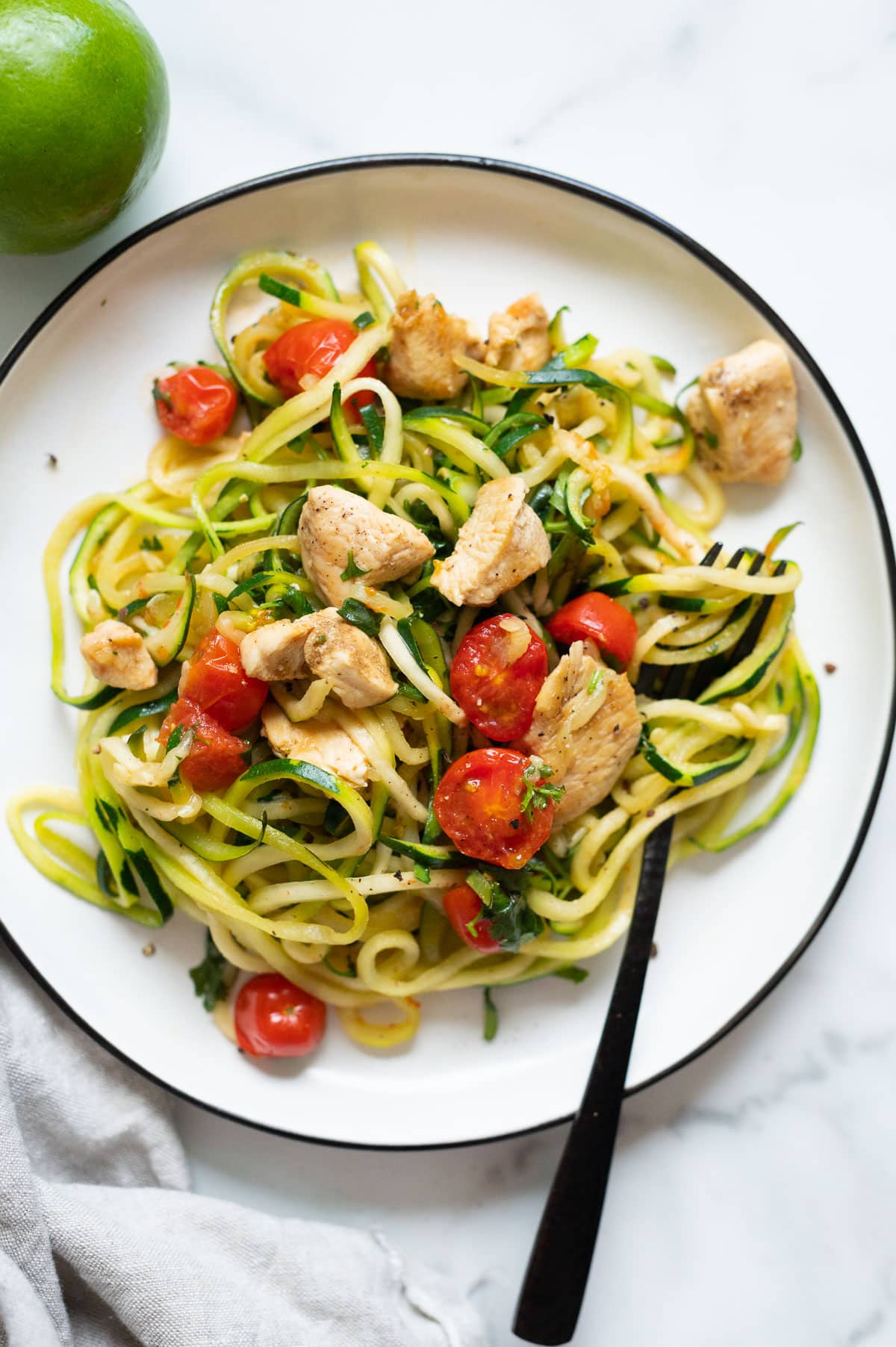 Chicken and zucchini noodles served on a plate with a fork. Lime and linen napkin on a countertop.