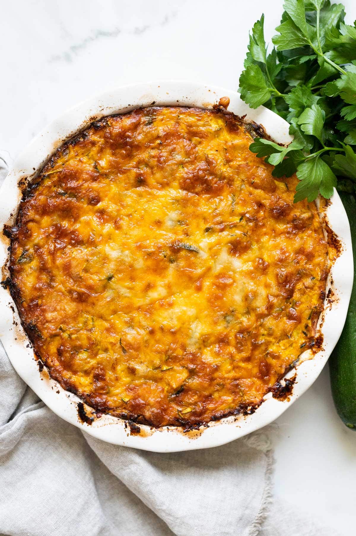 Crustless zucchini pie in a baking dish with fresh herbs and linen towel on a counter.