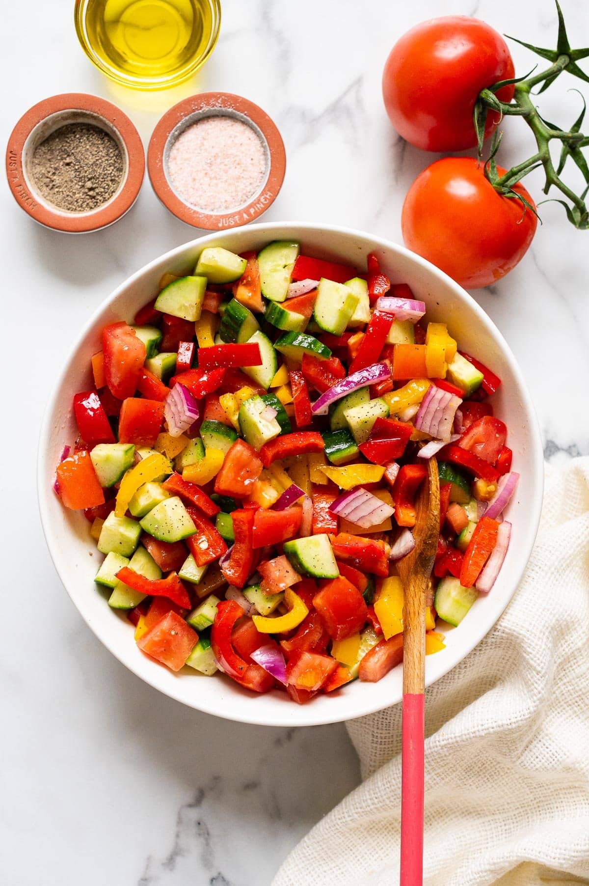 Tomato, cucumber bell pepper salad in white bowl with wooden spoon. Oil, salt, pepper, tomatoes on the vine and napkin on a counter.