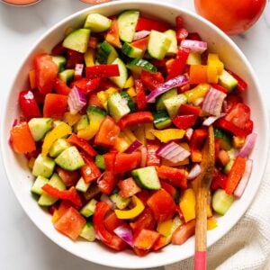 Tomato, cucumber bell pepper salad in white bowl with wooden spoon.