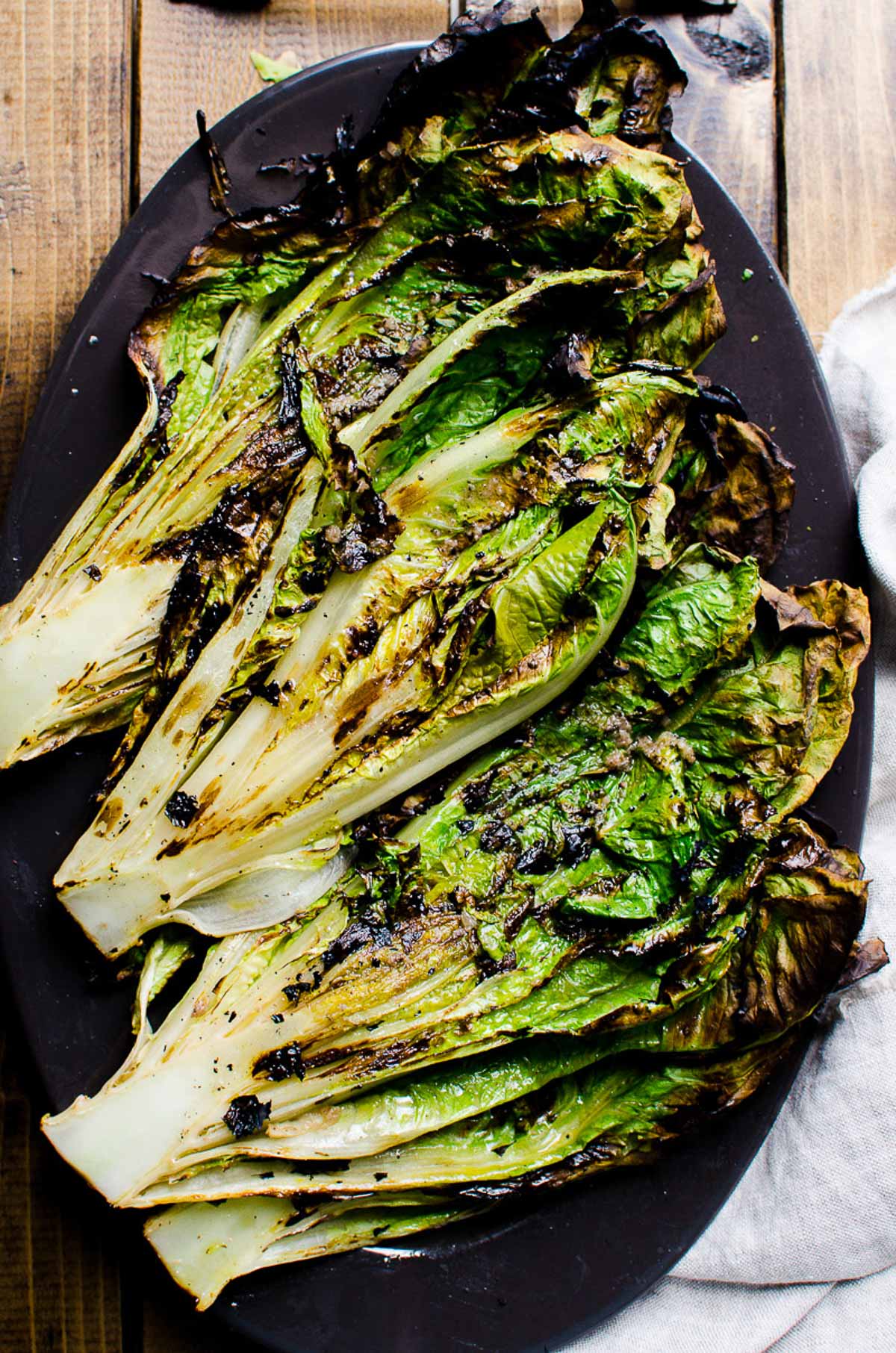 Grilled romaine lettuce served on black platter on wooden table. Linen napkin near it.