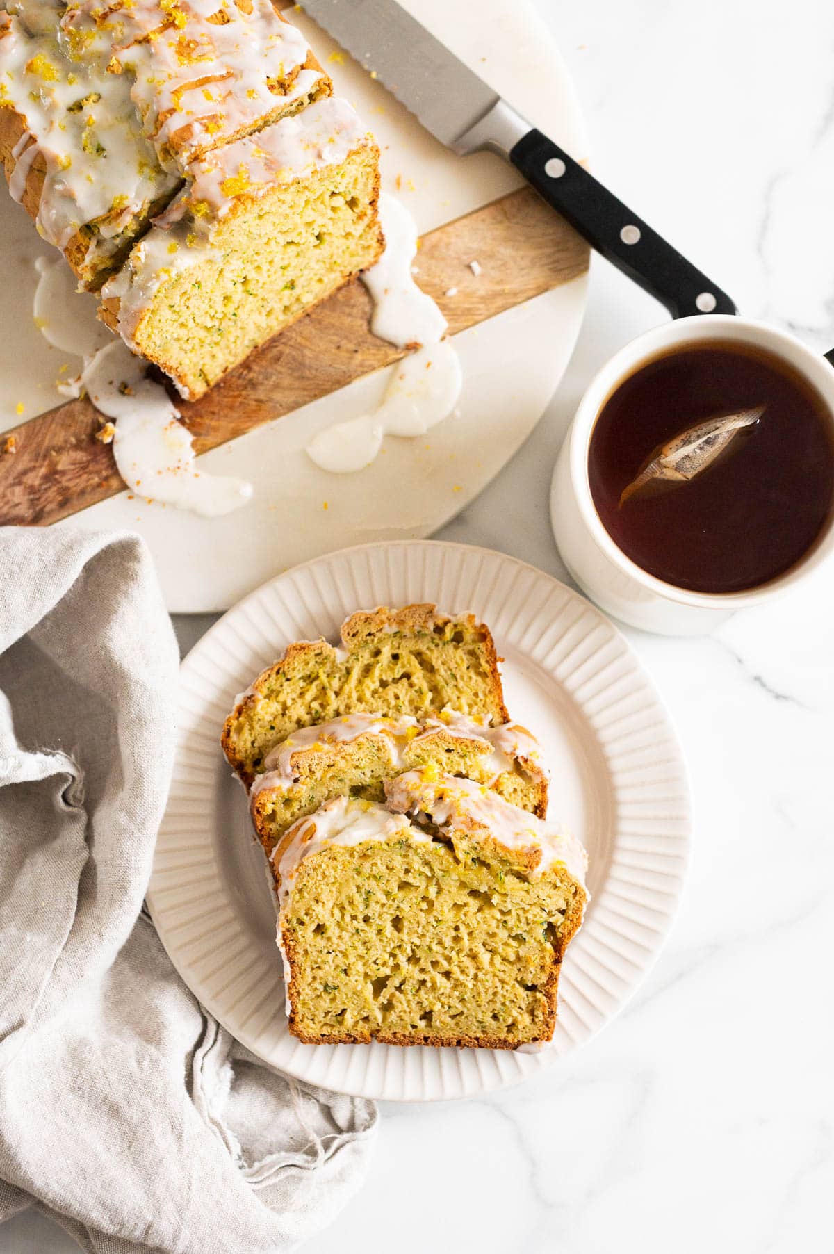 Lemon zucchini bread slices on a plate, lemon zucchini loaf on a platter and a cup of tea.