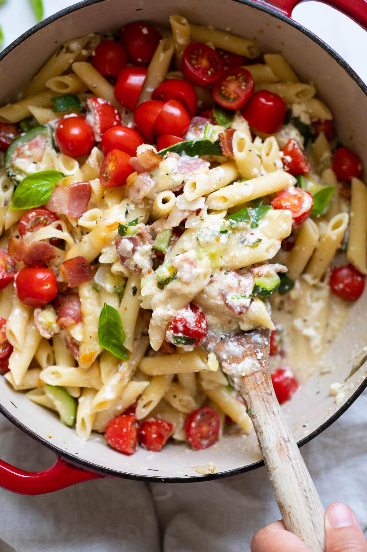 Close up of pasta with ricotta, bacon, zucchini, grape tomatoes, basil on a wooden spoon above the pot.
