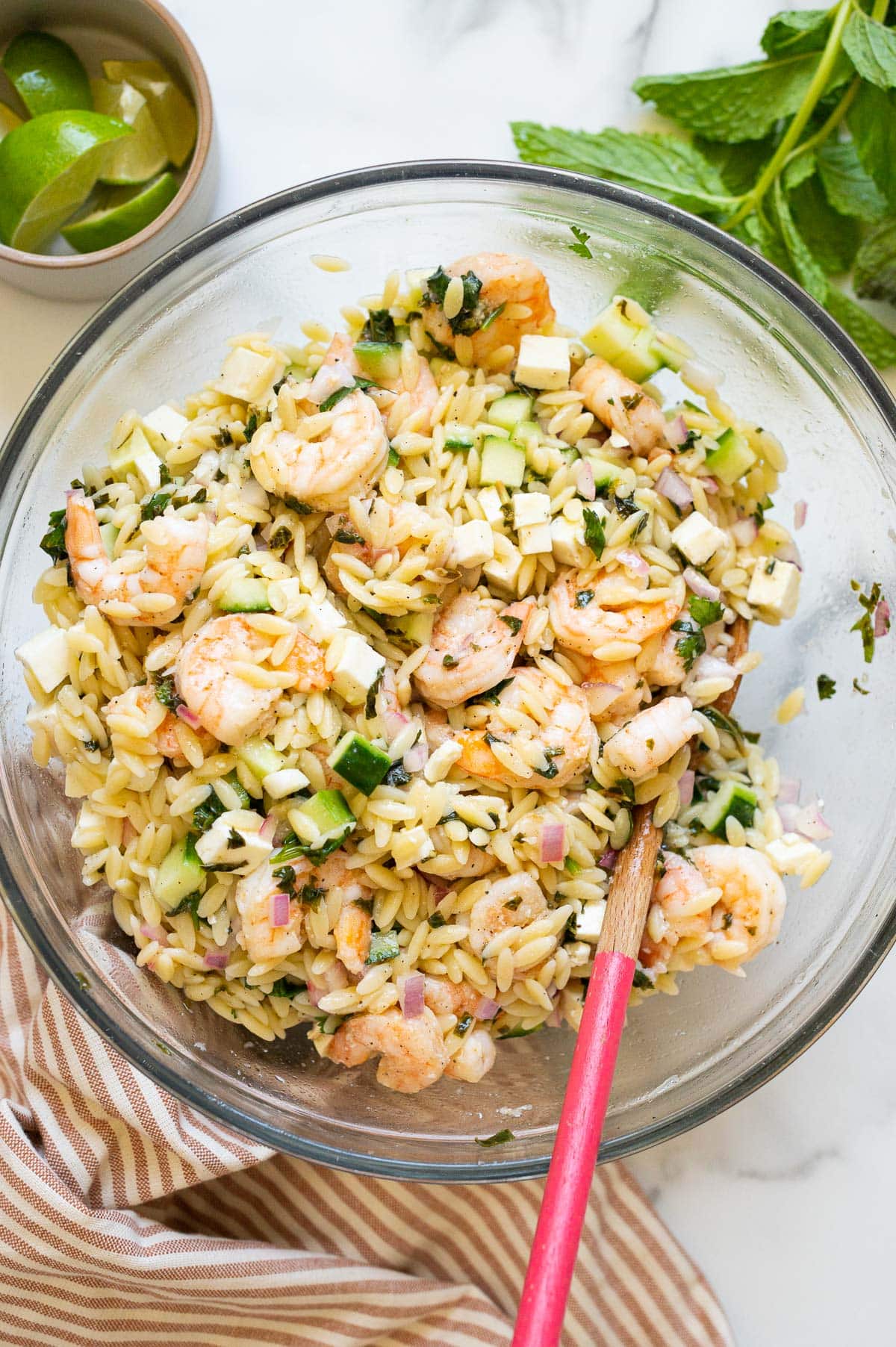Shrimp orzo salad with cucumbers in a bowl with wooden spoon. Lime, mint and napkin on a counter.