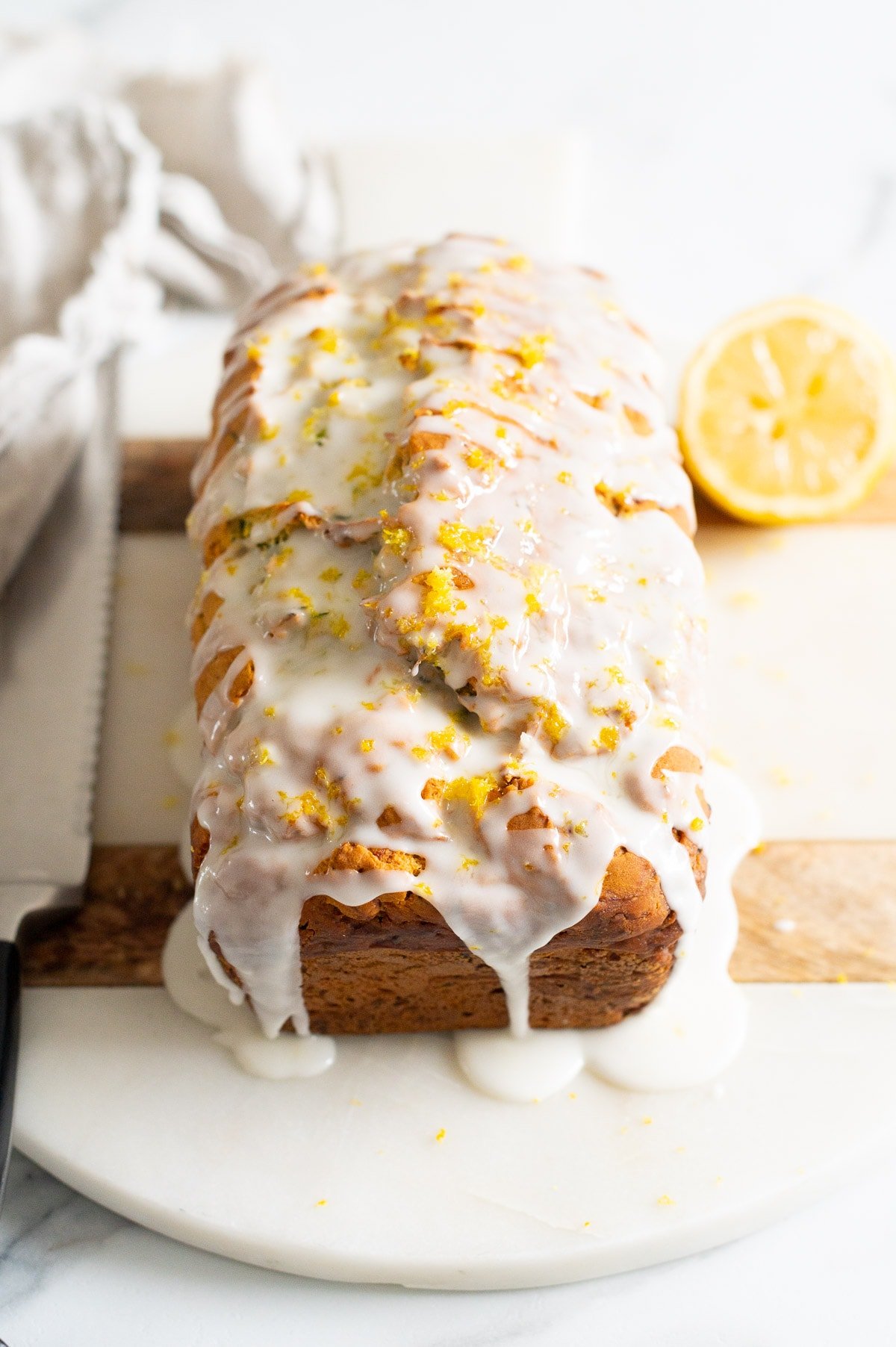 Lemon zucchini bread with icing and lemon zest on top served on a platter. Knife, linen napkin and lemon.