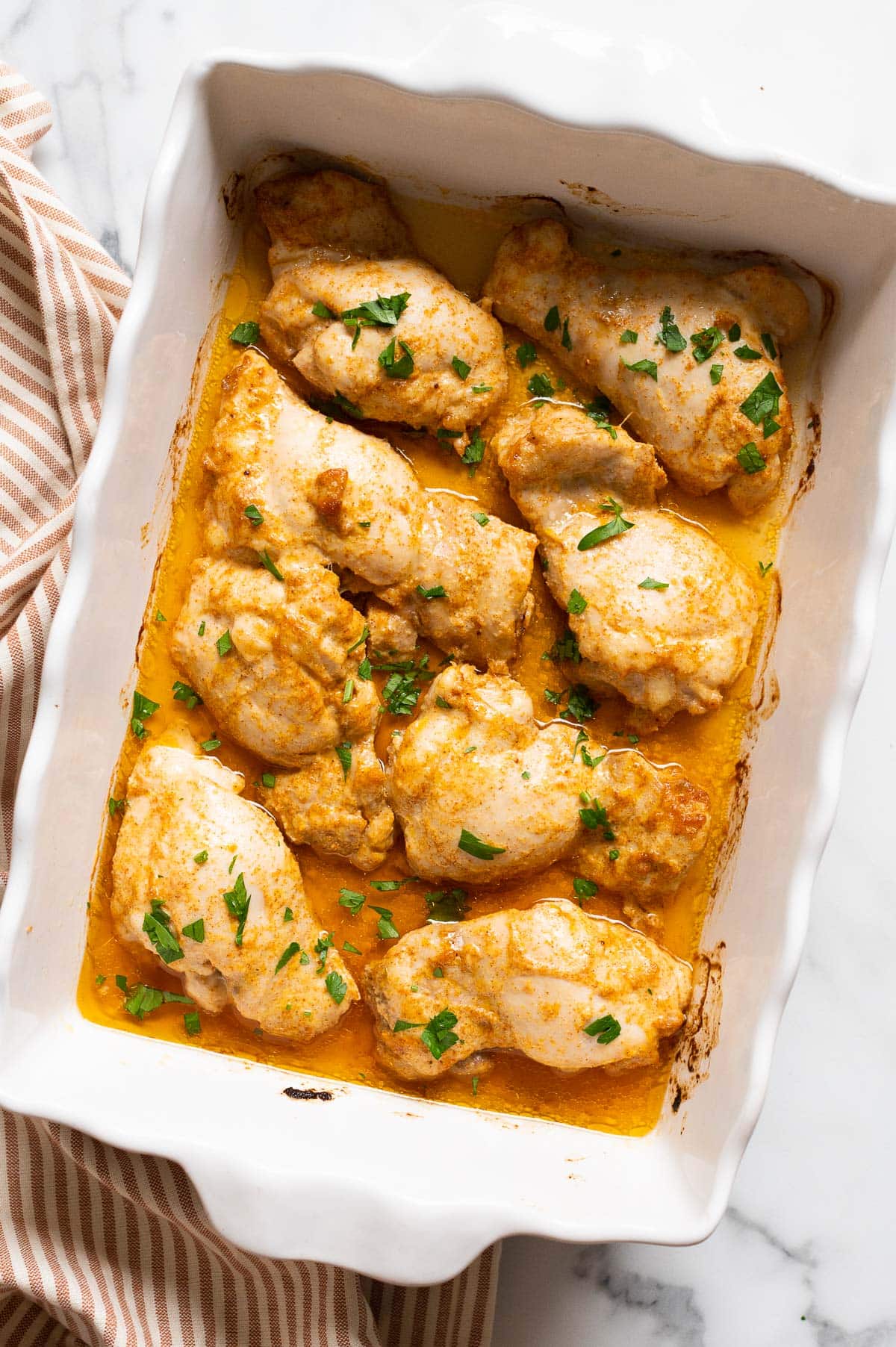 Eight baked boneless chicken thighs garnished with parsley and white dish. Linen towel on a counter.