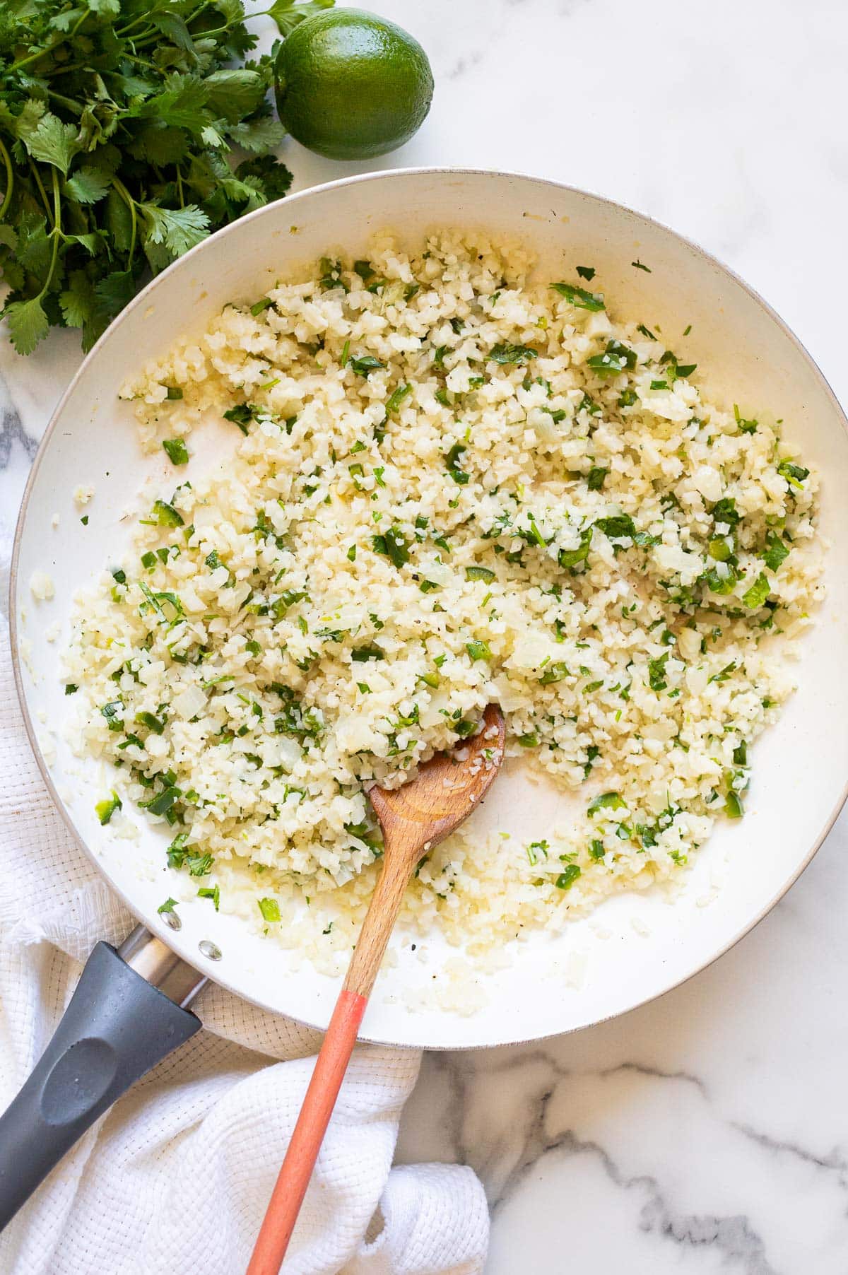 Cilantro lime cauliflower rice in a skillet with a wooden spoon. Bunch of cilantro, lime and linen napkin on a counter.