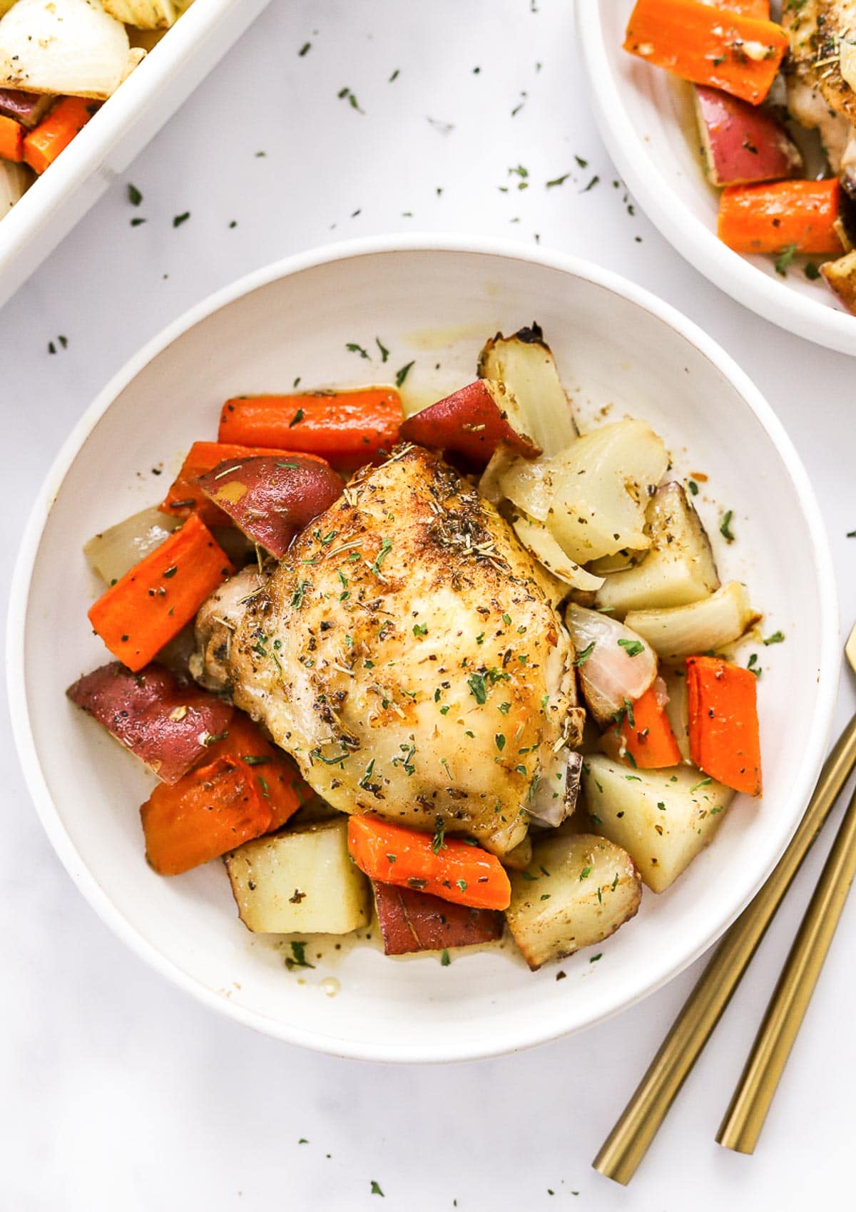 Roasted chicken thighs and potatoes with carrots served in a bowl and garnished with herbs.