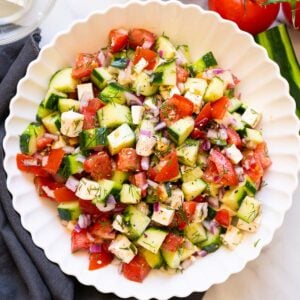 Cucumber tomato feta salad served in white bowl. Cucumbers, tomatoes and linen napkin on a counter.