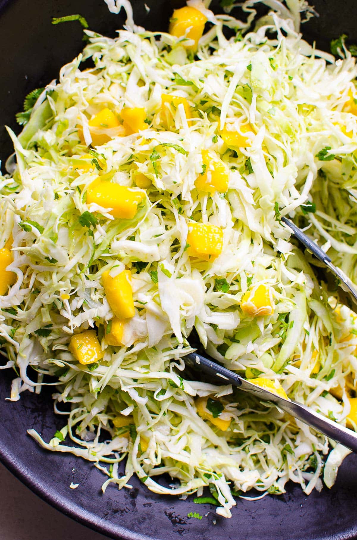 Mango slaw served in a black bowl with tongs.